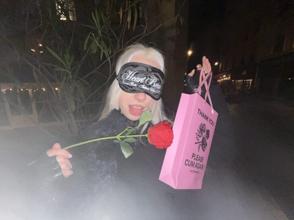 A person with platinum blonde hair wears a black sleep mask that says Heart Breaker, holds a red rose in their mouth, and carries a pink gift bag with black text, standing outdoors at night.