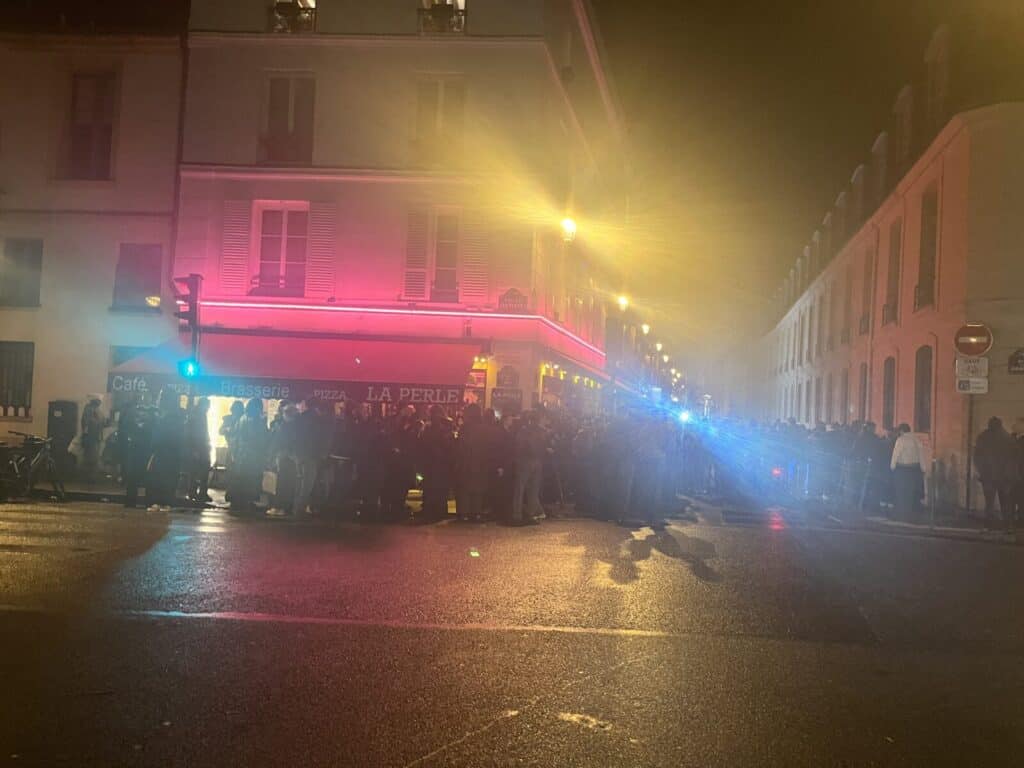 A crowd gathers on a wet street at night outside a building with pink neon lights. Police lights flash blue nearby, and the illuminated sign above the entrance reads La Perle.