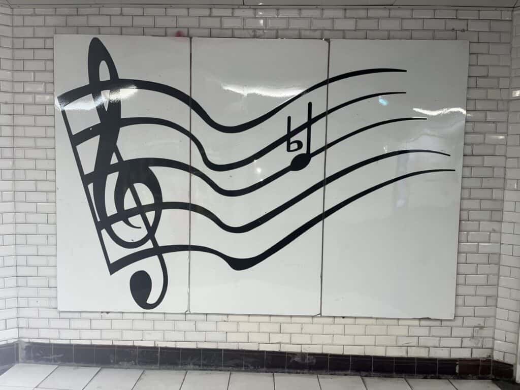 A mural on a white tiled wall shows a large black treble clef, musical staff, and a flat note, all painted in a bold, stylized design.