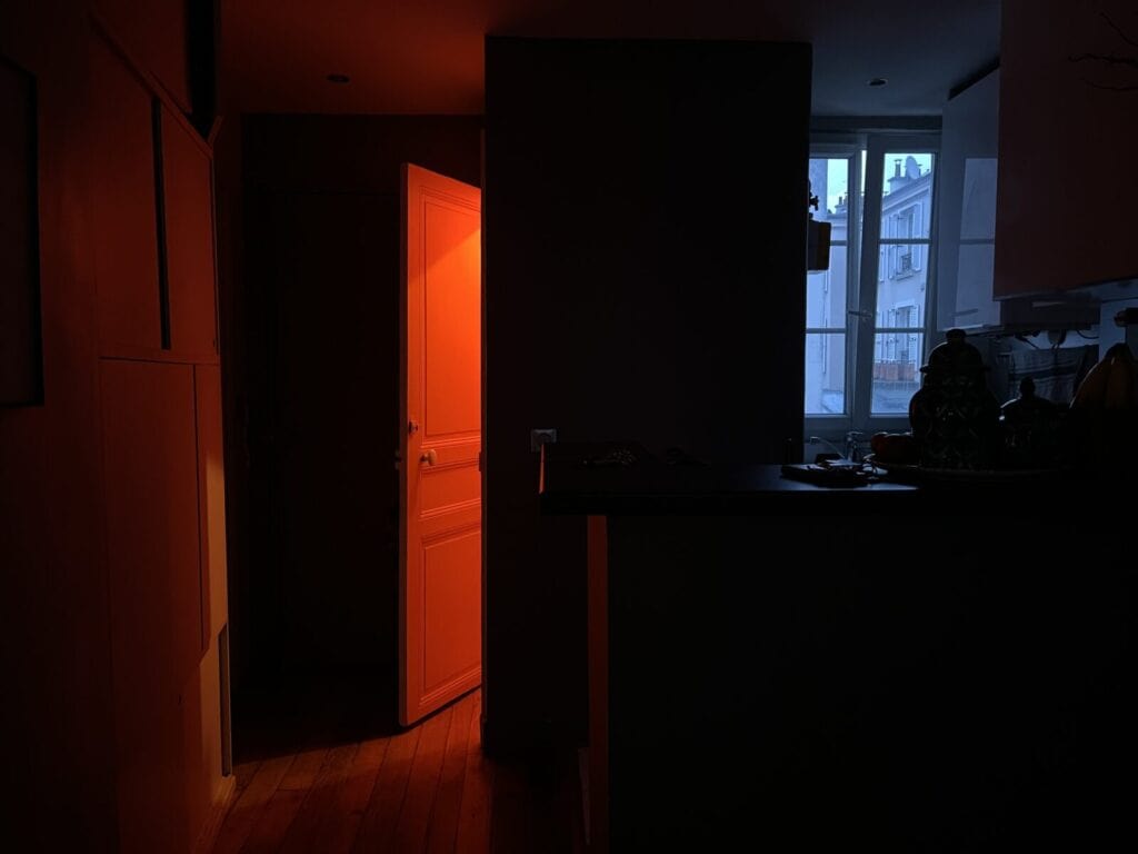 A dimly lit room with an open door glowing with warm orange light, contrasting with the cool blue daylight from a window in the kitchen area. Objects on the counter are in shadow.
