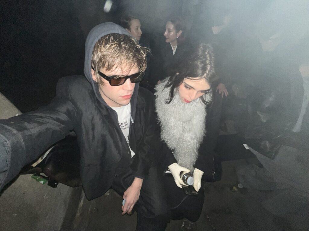 Two people sit close together in a dim, smoky setting. The person on the left wears sunglasses and a black hoodie, while the person on the right wears a fluffy scarf and white gloves, looking down at something in their hands. Other people are in the background.