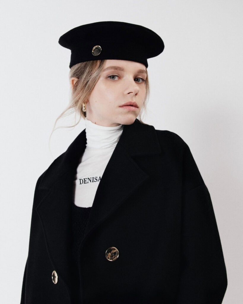 A woman wearing a black Denisa Rad coat with gold buttons, a white turtleneck, and a black beret with a gold pin stands against a plain white background, channeling the chic style of Milan Loves Seoul as she gazes slightly to the side.