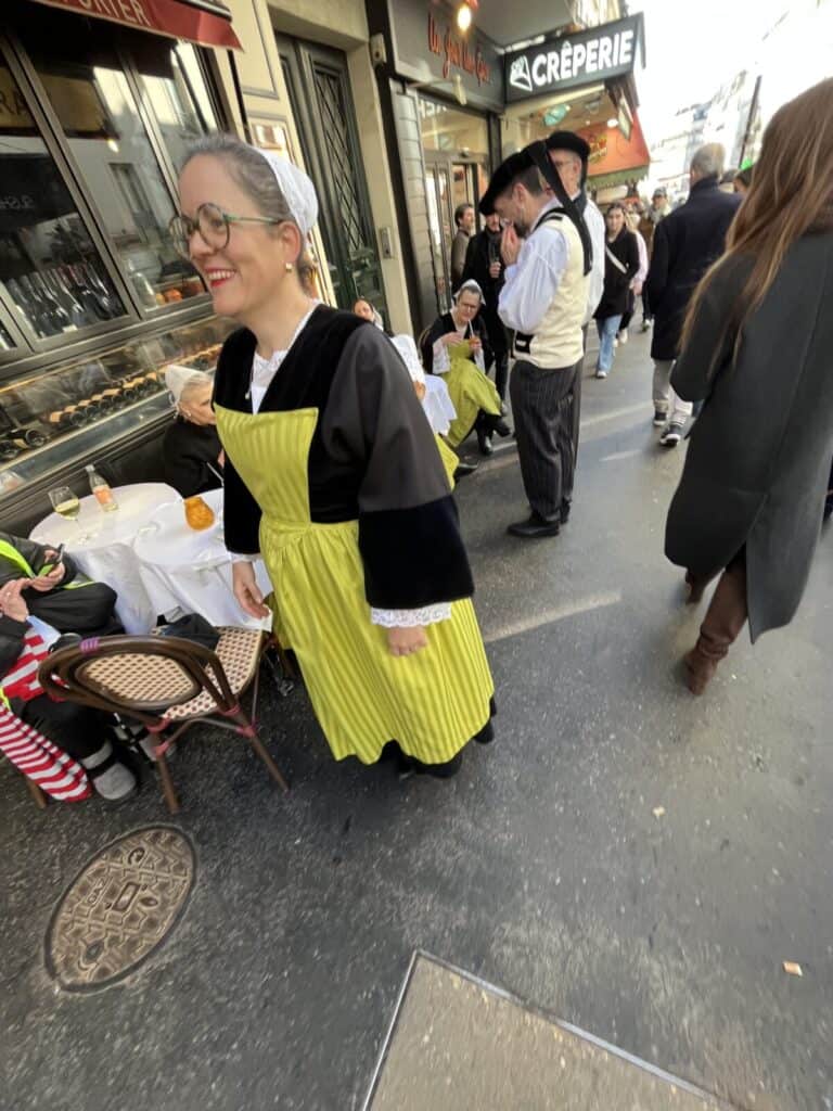 People in traditional attire, including a smiling woman in a yellow apron and white headscarf, stand on a busy Parisian sidewalk near outdoor café tables and a crêperie. Other pedestrians walk by.