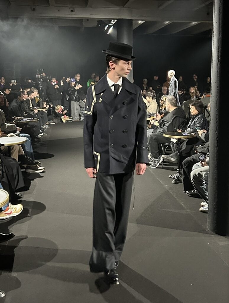 A male model in a black double-breasted coat with white trim and a black top hat walks a runway, surrounded by seated spectators watching a fashion show in a dimly lit venue.