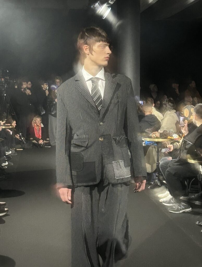 A male model walks a runway wearing a pinstripe suit with mismatched patchwork details, a white shirt, and a striped tie. The audience sits on either side, many holding phones and cameras, in a dimly lit fashion show setting.