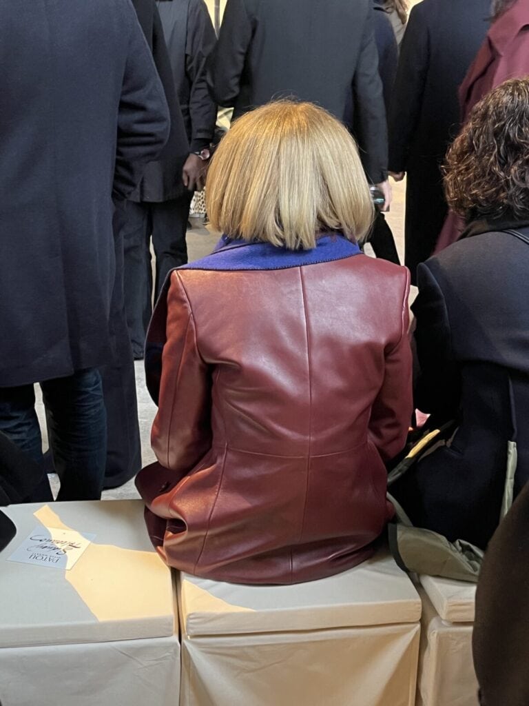 A person with a short blonde bob haircut, wearing a red leather coat with a blue collar, sits on a white cube seat among a group of people dressed mostly in dark clothing. The scene appears to be indoors.