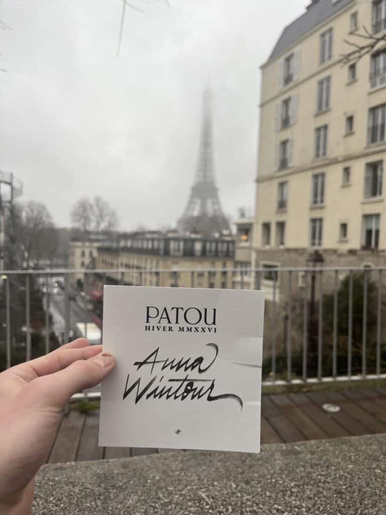 A hand holds a card reading PATOU HIVER MMXXVI Anna Wintour in front of a Parisian street with the Eiffel Tower visible in the foggy background.
