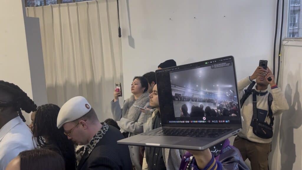 A person holds up a laptop displaying a live video of a crowded event, while people nearby use their phones to take photos or record. The scene appears busy, with attendees dressed in coats and hats.