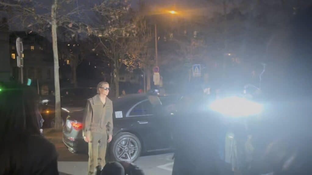 A person wearing sunglasses and a brown outfit stands on a street at night, facing bright camera flashes. Behind them, a black car is parked, and leafless trees and buildings are visible in the background.