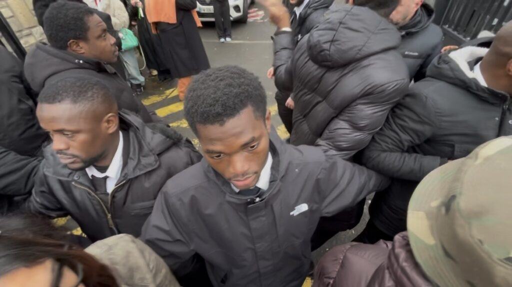 A group of people gather closely together outdoors, many wearing dark jackets. One man in the center looks down, while others around him face different directions, some with their backs to the camera.