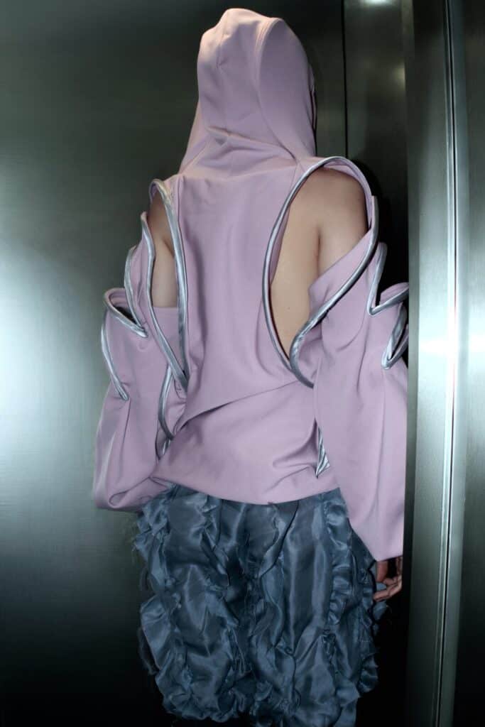 A person stands in an elevator, wearing a light pink hooded top with dramatic cut-out sleeves and shiny silver trim, paired with a ruffled, textured gray skirt. The person faces away, revealing their back and shoulder.