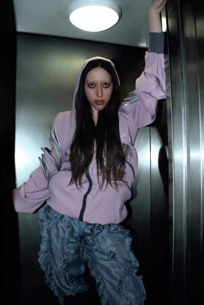 A person with long dark hair and bold makeup wears a light pink zip-up hoodie with metallic details and ruffled blue pants, posing confidently in a metallic elevator under a round ceiling light.