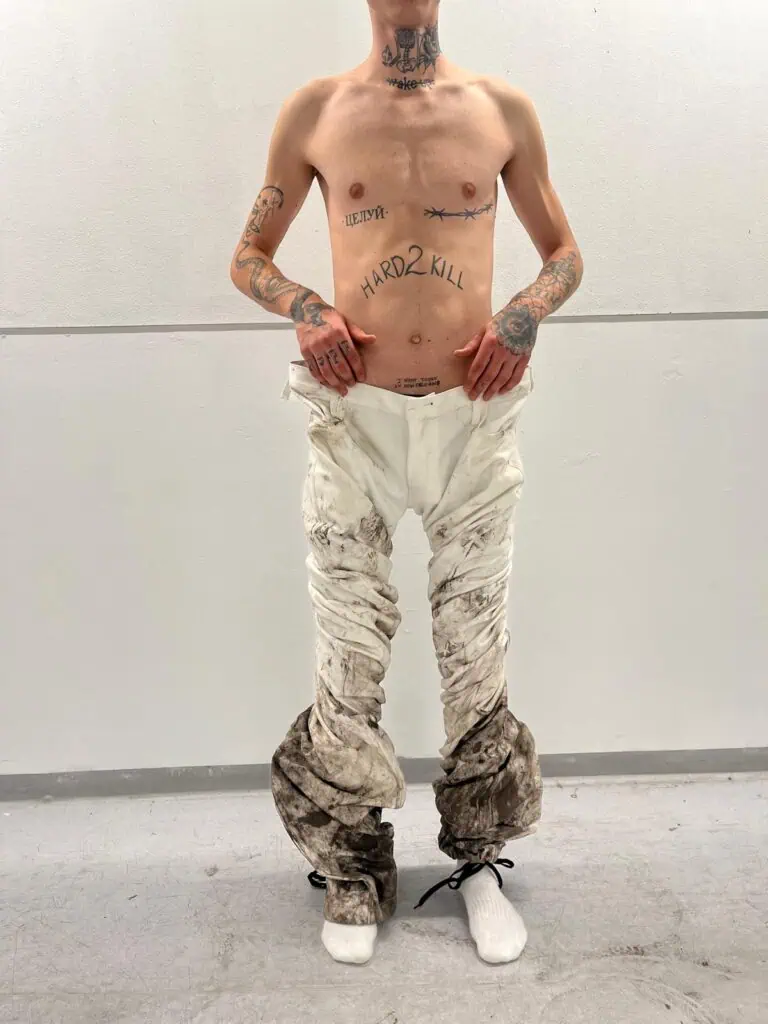 A shirtless person with tattoos on their chest, arms, and neck stands against a white wall —wearing loose, dirty white pants bunched by Kalina Dorożyńska Designs at the ankles and white socks; their hands rest on their hips.