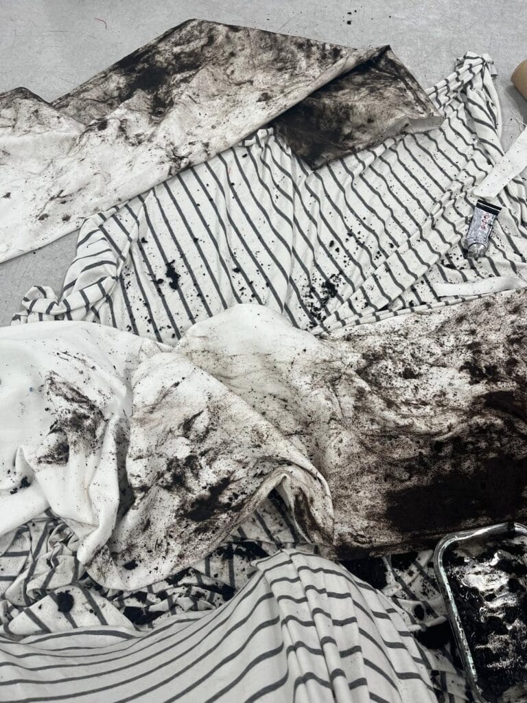 White and black striped fabric by Kalina Dorożyńska Designs is sprawled on the floor, heavily smeared and splattered with black dirt or paint. Among the messy fabric, a black and white tube of paint and an open tray with dark liquid evoke a Ben Kreukniet-inspired scene.