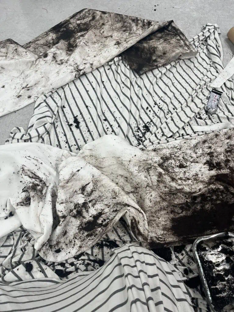 White and black striped fabric by Kalina Dorożyńska Designs is sprawled on the floor, heavily smeared and splattered with black dirt or paint. Among the messy fabric, a black and white tube of paint and an open tray with dark liquid evoke a Ben Kreukniet-inspired scene.