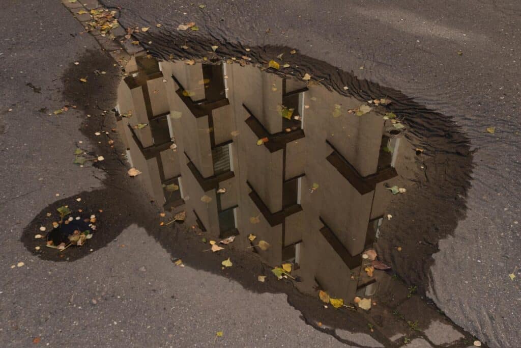 A puddle on an asphalt street reflects the facade and windows of a building, surrounded by scattered fallen leaves and a nearby manhole cover. Image by Kalina Dorożyńska.