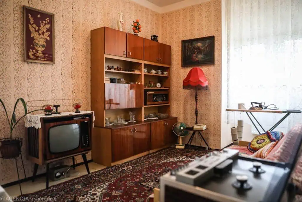A vintage living room with patterned wallpaper, an old TV, wooden cabinet, red lampshade, radio, fan, iron board, and a patterned rug evokes a retro 1970s or 1980s atmosphere reminiscent of Ben Kreukniet’s nostalgic style, image by Kalina Dorożyńska.
