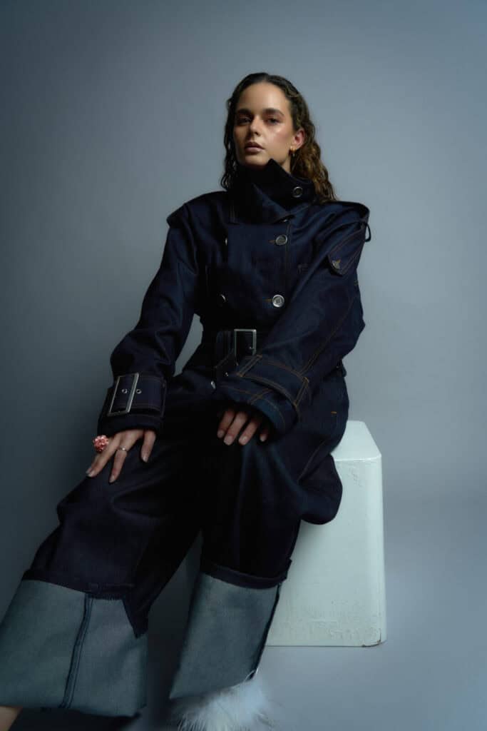A woman with wavy hair sits on a white block, wearing a dark, structured denim jumpsuit with wide cuffs and oversized sleeves. She looks confidently at the camera against a gray studio backdrop.