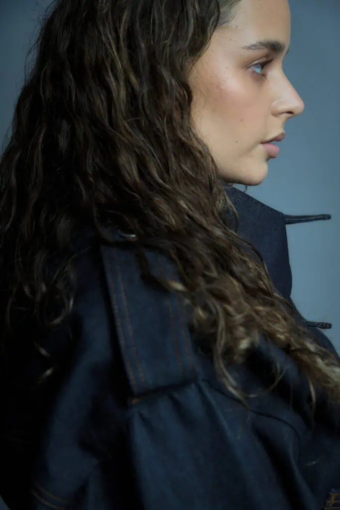 A woman with long, wavy brown hair is shown in profile against a gray background, wearing a dark denim jacket with visible stitching. Her expression is calm and contemplative.