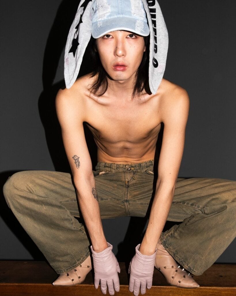 A shirtless person squats on a wooden surface, wearing brown pants, beige gloves, and a light denim cap with floppy bunny ears. The background is dark, creating a dramatic and edgy atmosphere.