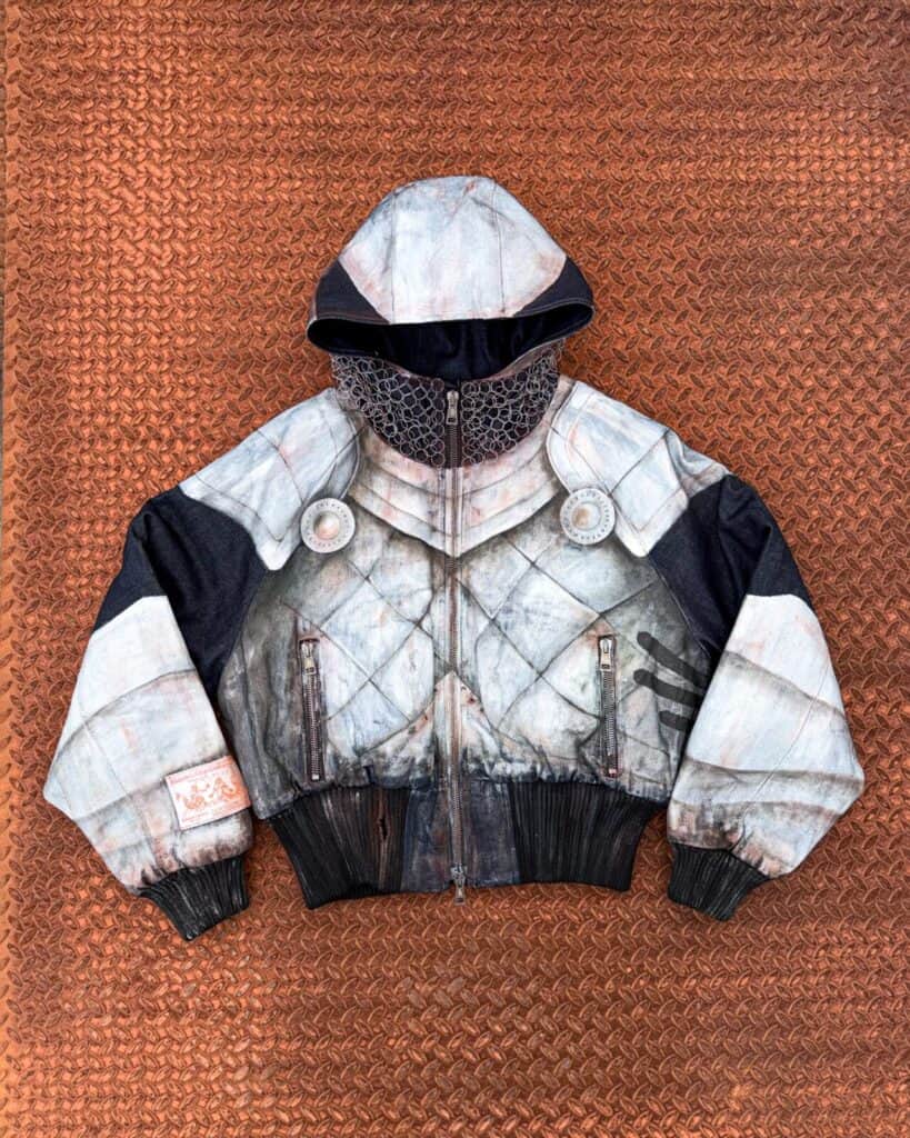 A hooded jacket with a detailed, realistic print resembling medieval armor lies flat on a rust-colored, diamond-patterned metal surface. The design includes metallic textures, rivets, and a chainmail-style hood.