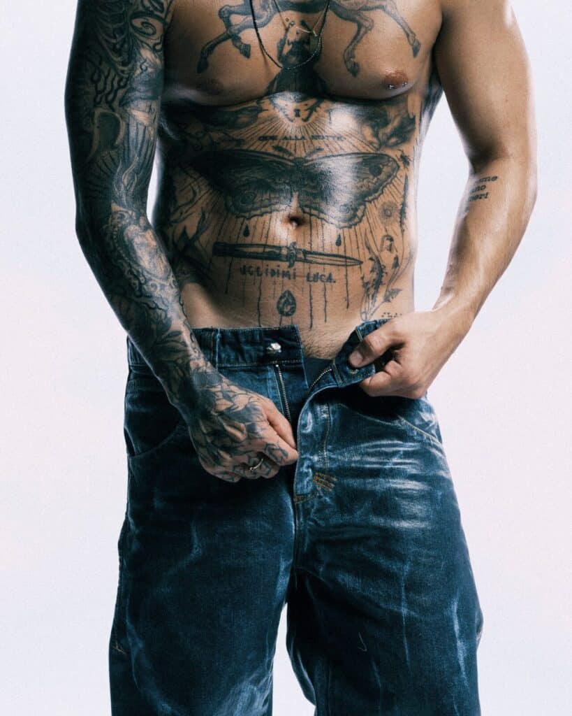 A shirtless person with extensive tattoos on their chest, arms, and torso is standing and pulling up blue jeans, partially unzipped, against a plain light background.