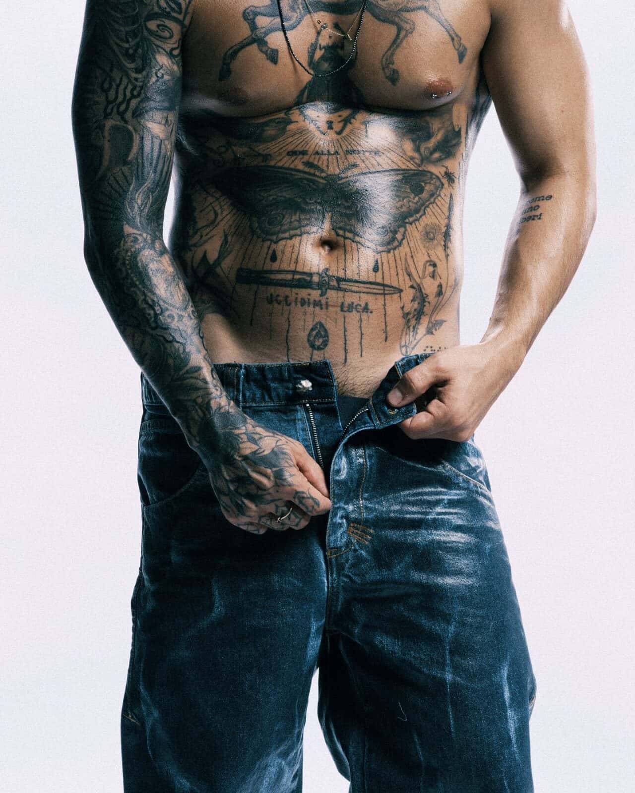 A shirtless person with extensive tattoos on their chest, arms, and torso is standing and pulling up blue jeans, partially unzipped, against a plain light background.
