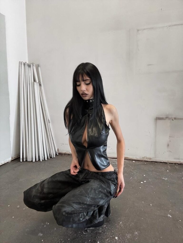 A person with long black hair, wearing a sleeveless black leather top and black pants, kneels on a concrete floor in a minimalist, industrial-looking room with white pipes stacked against the wall.