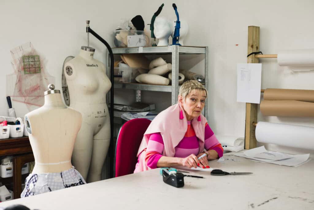 A woman wearing a pink striped sweater sits at a workspace with sewing tools and fabric. Behind her are mannequins, shelves with rolls of material, and various sewing supplies. She looks thoughtfully towards the camera.