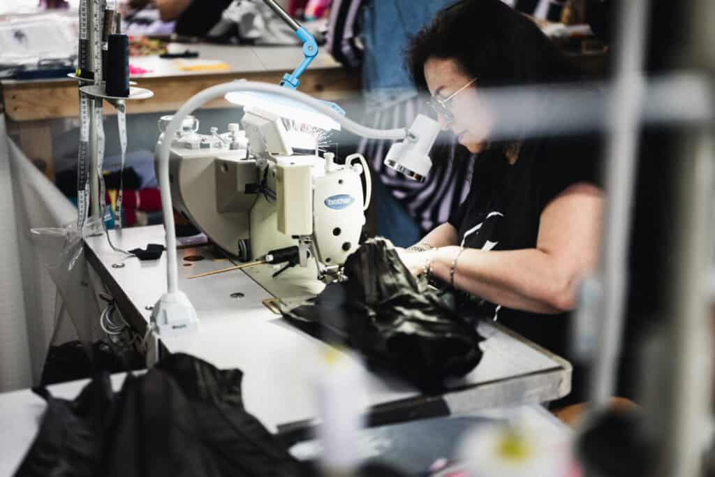 A woman with long dark hair and glasses operates a sewing machine, working on a piece of black fabric in a well-lit workshop, surrounded by sewing tools and materials.