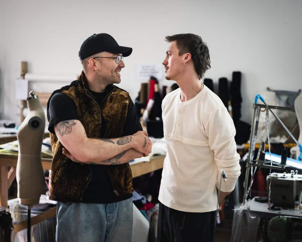 Two men stand and talk in a workshop or studio. One wears a black cap, glasses, and a brown vest with visible tattoos on his arms; the other wears a white shirt. Sewing equipment and mannequins are in the background.
