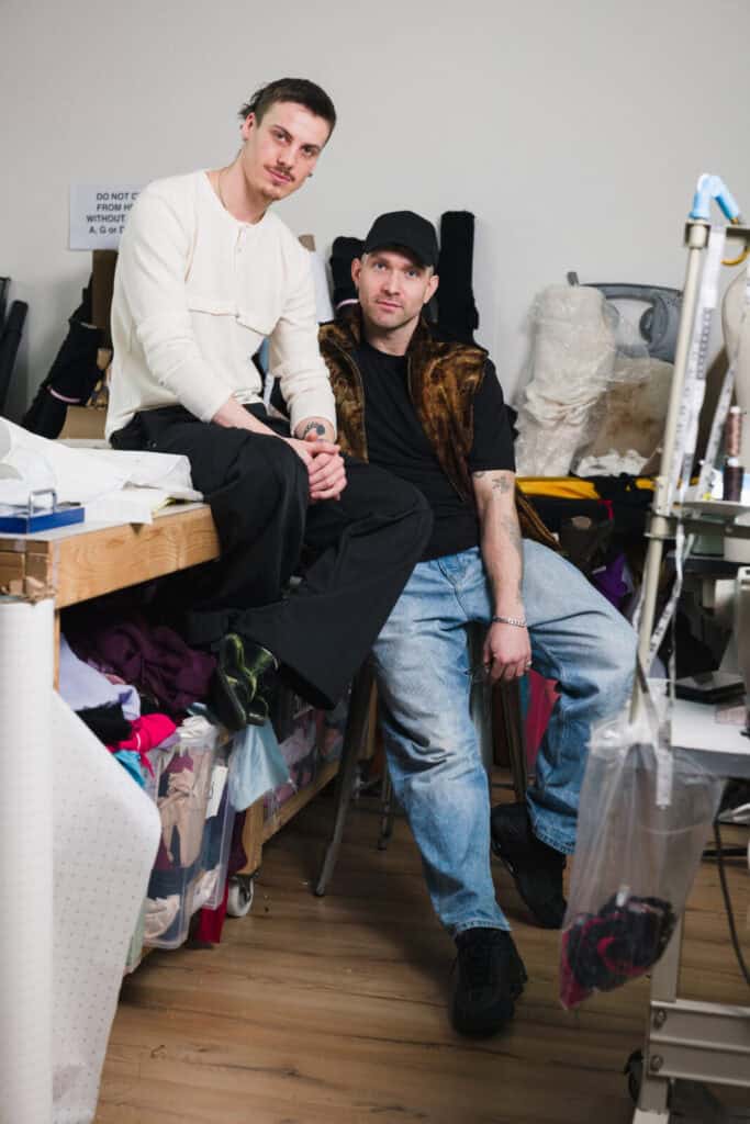 Two men pose in a fashion studio filled with fabric and sewing supplies. One man sits on a table while the other sits on a stool beside him. Both look at the camera and are dressed casually in modern clothing.