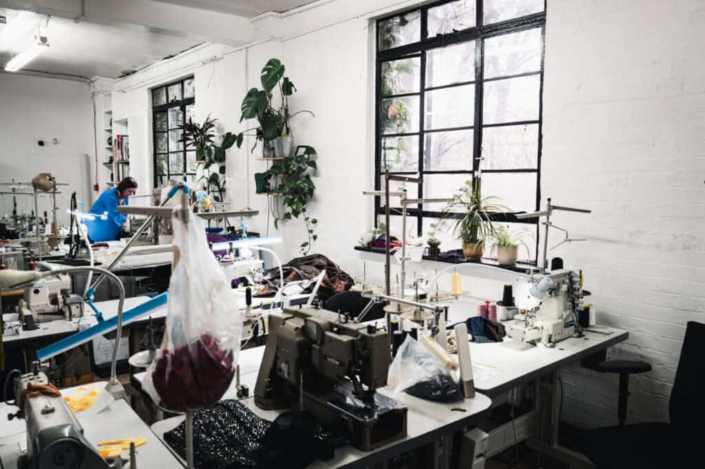 Bright sewing studio with several sewing machines on tables, potted plants on windowsills, and two people working in the background near large windows letting in natural light.