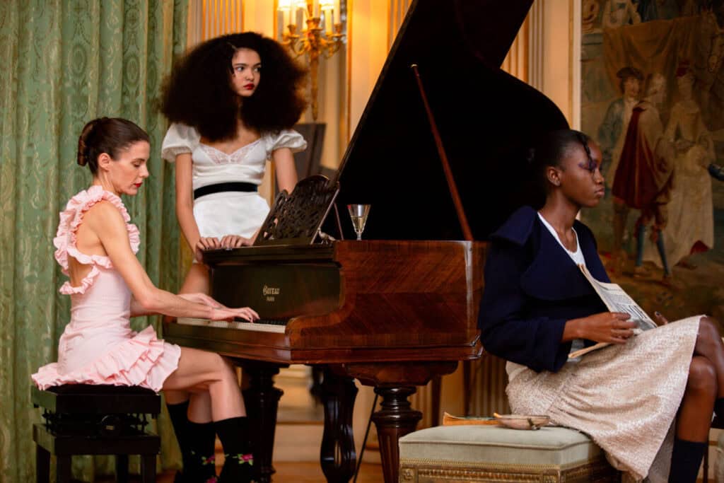 Three women are in an elegant room by a grand piano; one in a pink ruffled dress plays the piano, another in a white dress stands behind, and a third in a navy jacket and skirt sits holding sheet music.
