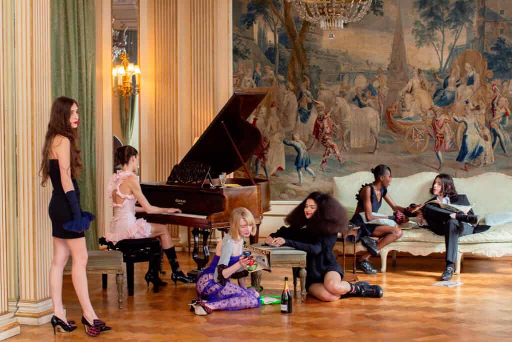 Six women in stylish, modern outfits socialize in an ornate room with a grand piano, classical columns, and a large tapestry. Some are sitting or reclining, while one stands by, and one plays the piano.
