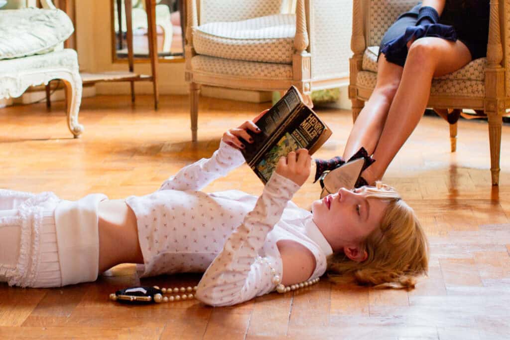 A person with light hair lies on a wooden floor reading a book, dressed in a white outfit and pearls, while another person sits nearby on an ornate chair with crossed legs, their face out of frame.