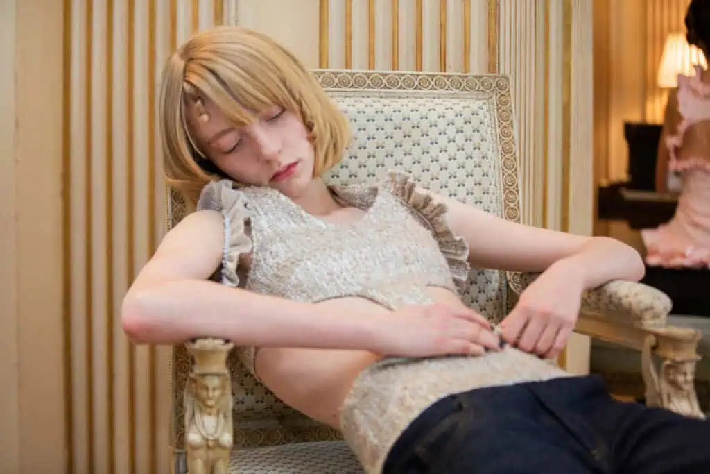 A person with short blond hair wearing a sleeveless knit top and jeans is reclining with eyes closed in an ornate armchair, appearing relaxed or sleepy, in an elegant room with patterned walls and soft lighting.