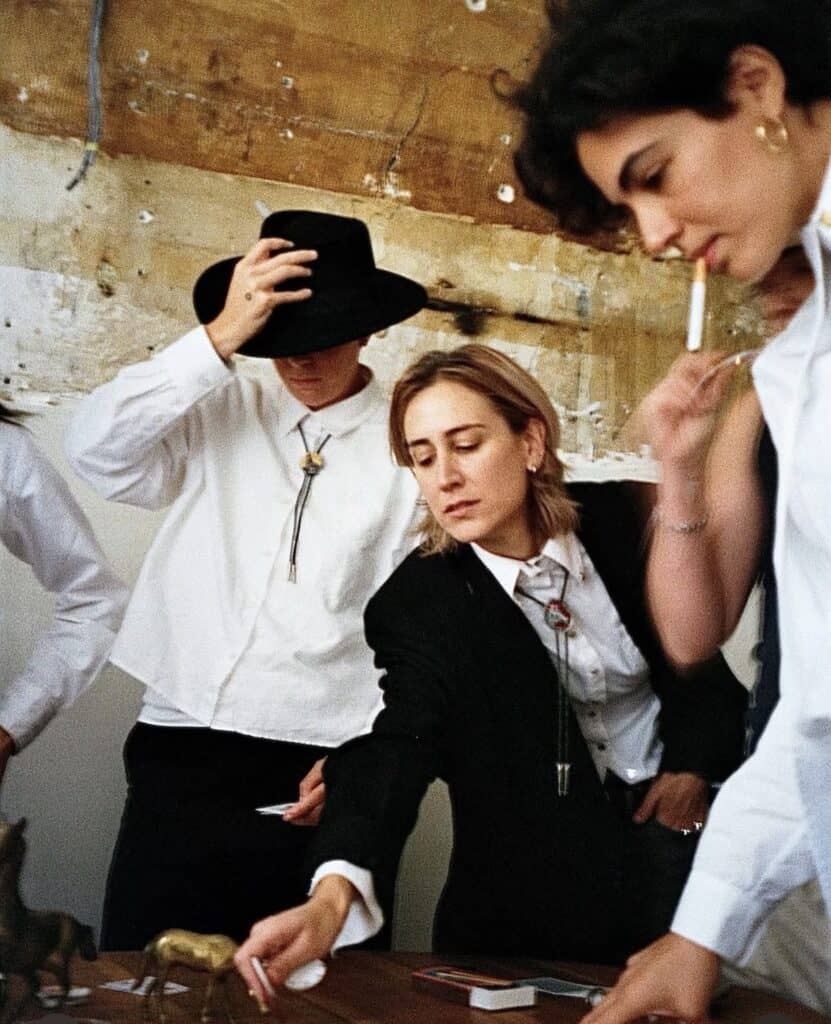 Three people in suits stand around a table. One wears a cowboy hat and holds it down, another leans forward to reach for something, and the third is standing, holding a cigarette. The atmosphere is casual and focused.