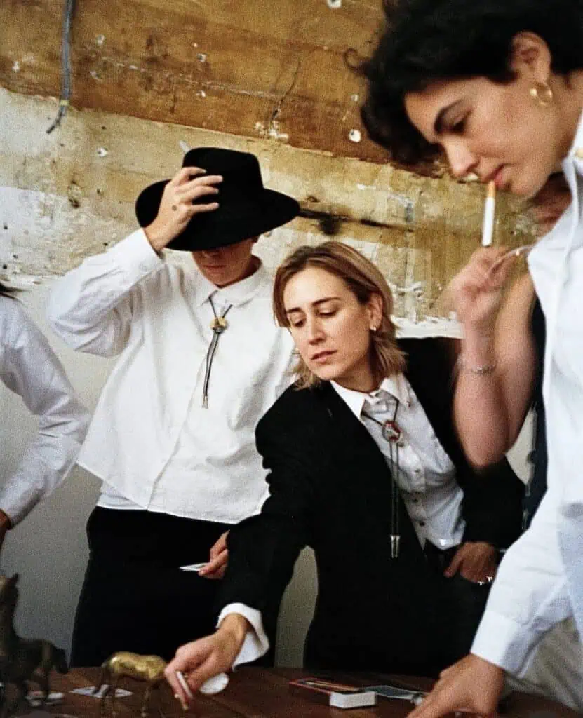 Three people in suits stand around a table. One wears a cowboy hat and holds it down, another leans forward to reach for something, and the third is standing, holding a cigarette. The atmosphere is casual and focused.