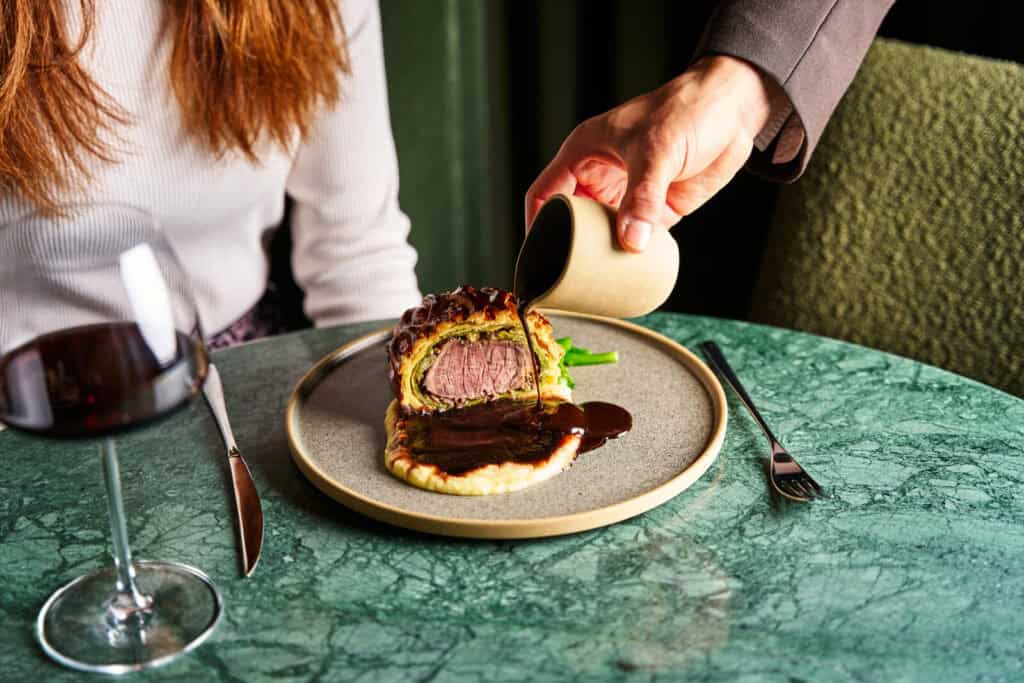 A person pours dark sauce over a beef Wellington served on a plate with mashed potatoes and greens, while a woman in a white sweater sits at a green marble table with a glass of red wine.