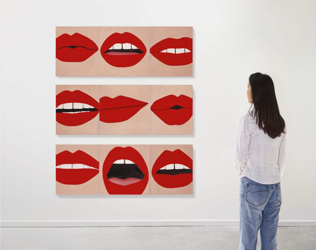 A woman stands in front of a wall displaying nine paintings of red lips in various shapes and expressions, arranged in three horizontal rows.