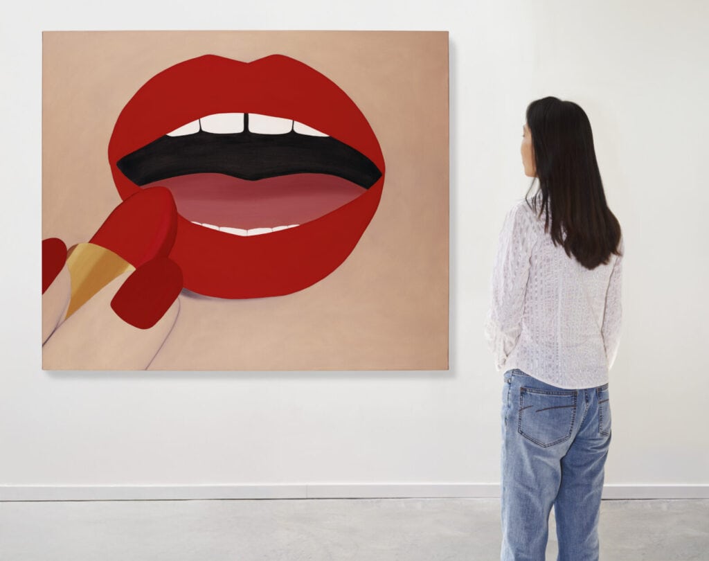 A woman in casual clothes stands in a gallery, looking at a large painting of red lips being colored with red lipstick on a beige background.