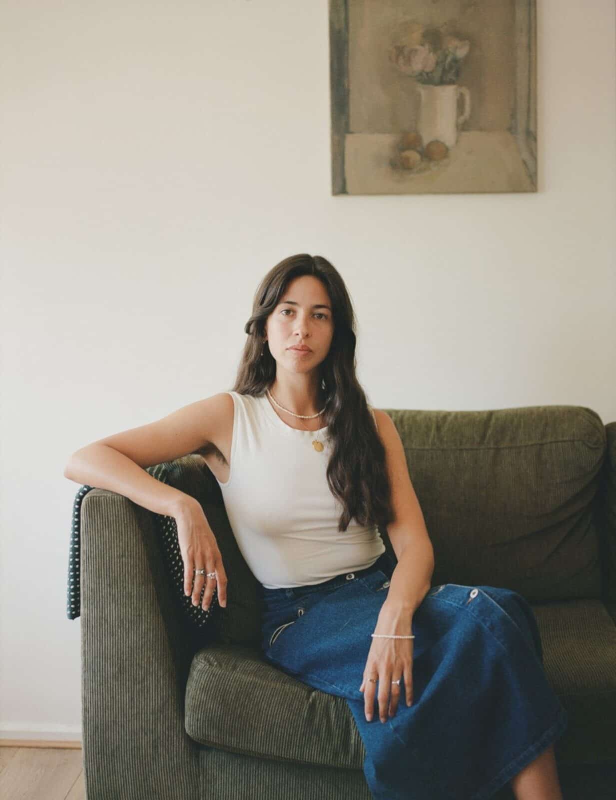 A woman with long dark hair sits on a green sofa, wearing a white sleeveless top and a blue skirt. She looks at the camera with a neutral expression. A painting of flowers in a vase hangs on the wall behind her.