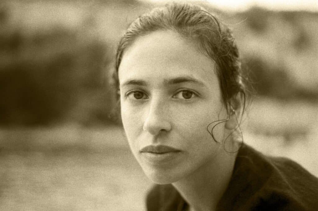 A sepia-toned close-up photo of a young woman with wavy hair pulled back, looking directly at the camera with a calm expression. The background is softly blurred, suggesting an outdoor setting.