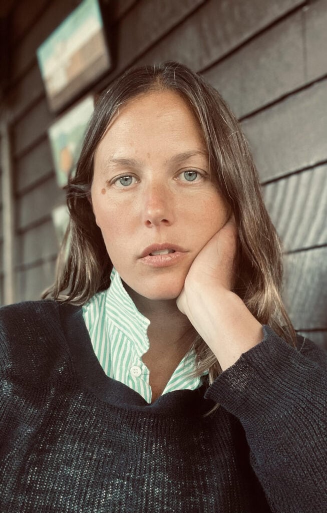 A woman with light skin and straight brown hair rests her face on her hand, wearing a striped shirt and dark sweater, sitting indoors against a wooden-paneled wall with framed pictures in the background.