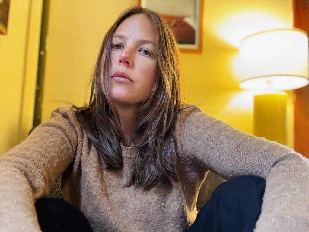 A woman with long brown hair wearing a beige sweater sits indoors with her knees up, looking at the camera. The background features a lit lamp and yellow walls with framed art.