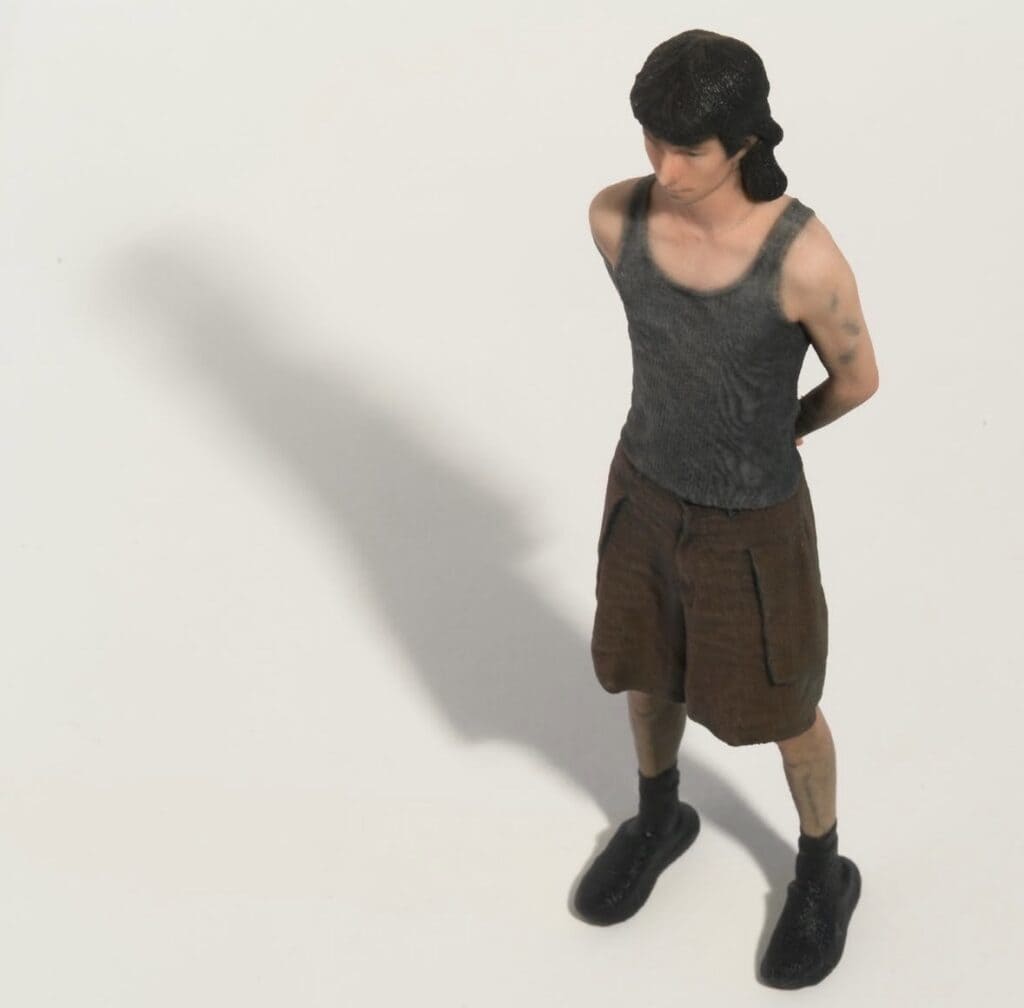 A small figurine of a person with short dark hair, wearing a gray tank top, brown shorts, and black shoes, stands with hands behind their back on a plain white surface, casting a shadow to the left.