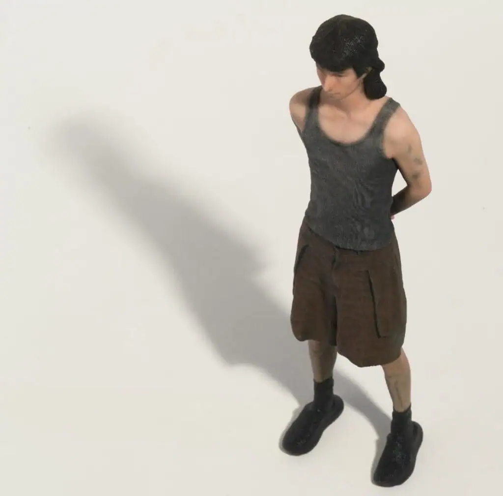 A small figurine of a person with short dark hair, wearing a gray tank top, brown shorts, and black shoes, stands with hands behind their back on a plain white surface, casting a shadow to the left.