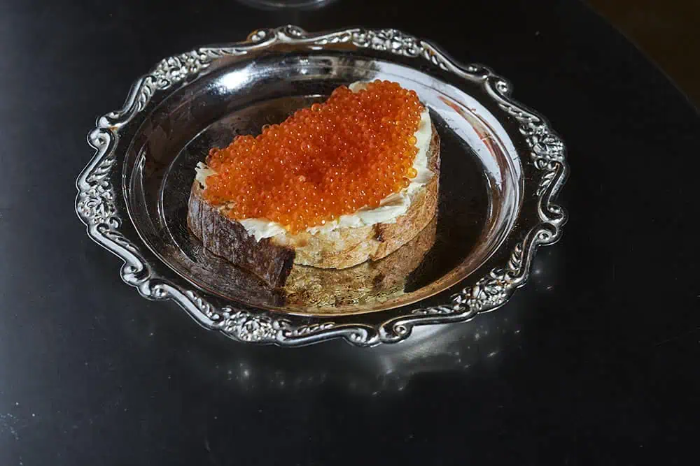 A slice of bread with butter and red caviar sits on an ornate silver plate, placed on a dark reflective surface—a decadent option for those seeking inspiration on where to eat during London Fashion Week.