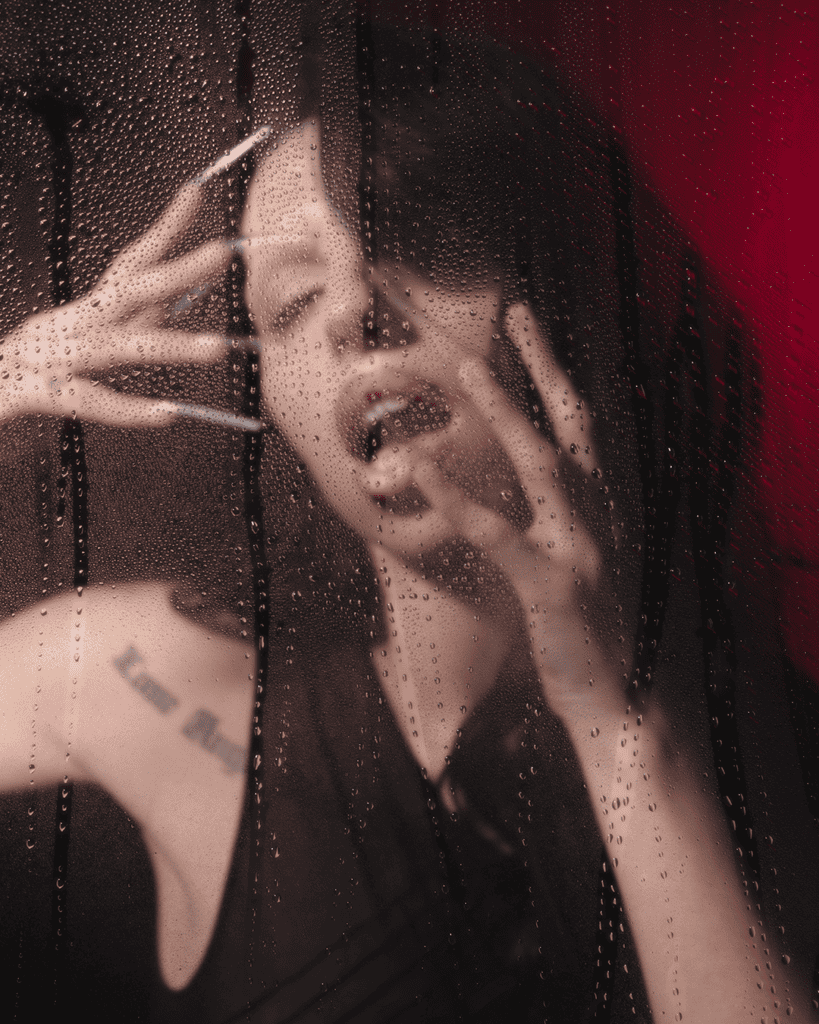 A woman with long nails poses dramatically behind a rain-streaked glass pane, with her mouth slightly open and hands pressed against the glass. The background is dark with hints of red light.